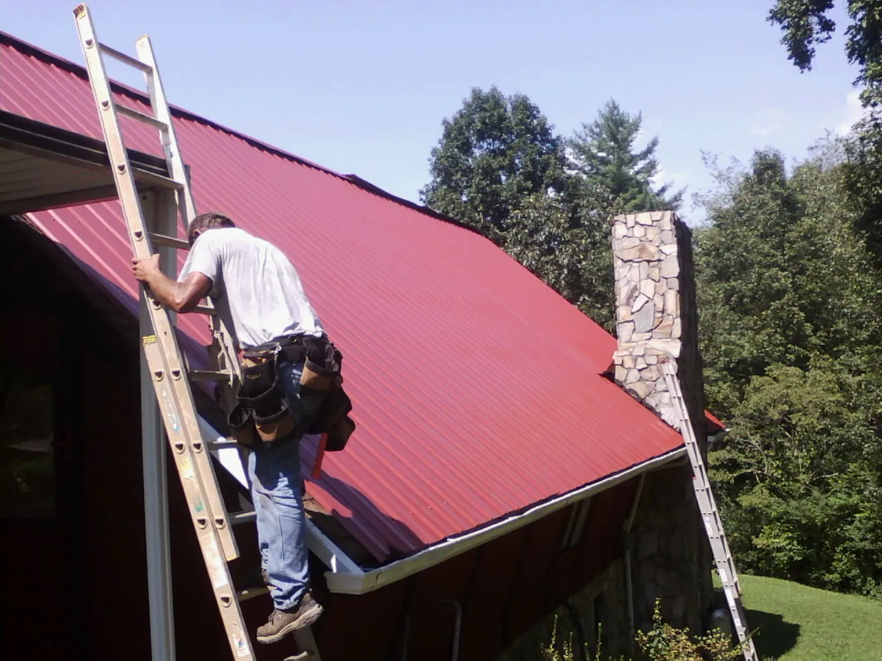 Roofer on a ladder beside a red metal roof and stone chimney — Soffit & Fascia Repair by Roofing Brothers in Mattapoisett
